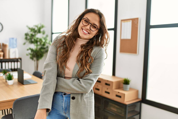 Молодая красивая латиноамериканка-предприниматель улыбается уверенно стоя со скрещенными руками в офисе - Фото, изображение