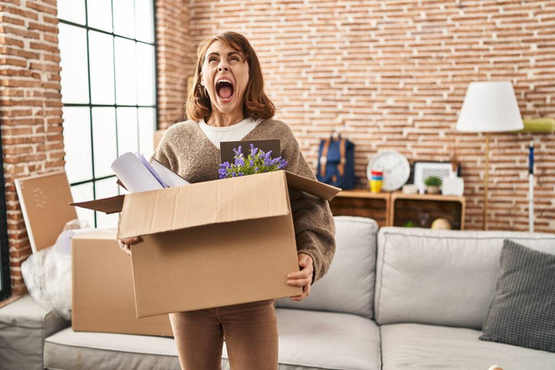 Jonge mooie vrouw holding box verhuizen naar een nieuw huis boos en gek schreeuwen gefrustreerd en woedend, schreeuwen van woede op zoek naar.  - Foto, afbeelding