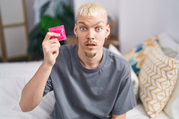 Jonge blanke man met condoom zittend op bed bang en verbaasd met open mond voor verrassing, ongeloof gezicht  - Foto, afbeelding