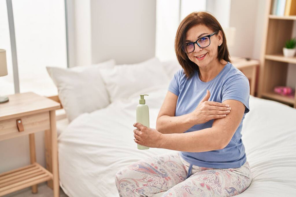 Middle age woman applying skin treatment sitting on bed at bedroom - Photo, Image