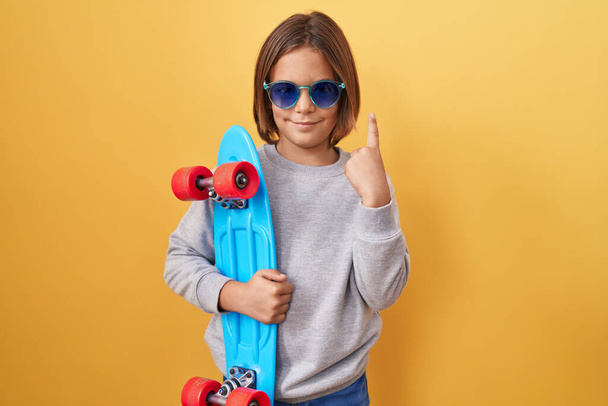 Pequeño niño hispano con gafas de sol sosteniendo patín sorprendido con una idea o pregunta apuntando con el dedo con la cara feliz, número uno  - Foto, Imagen