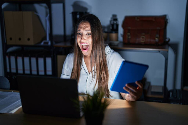 Young brunette woman working at the office at night angry and mad screaming frustrated and furious, shouting with anger. rage and aggressive concept.  - Photo, Image