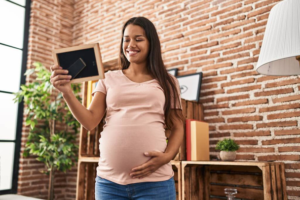 Mujer latina joven embarazada mirando foto en casa - Foto, imagen