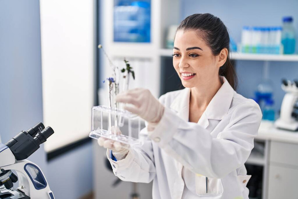 Young beautiful hispanic woman scientist holding test tubes with flowers at laboratory - Zdjęcie, obraz