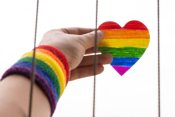 LGBTの虹の手首バンドを持つ男とワイヤーフェンスから紙の心を持ち上げる - 写真・画像
