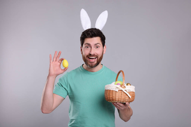 Portrait of happy man in cute bunny ears headband holding wicker basket with Easter eggs on light grey background - Foto, Bild