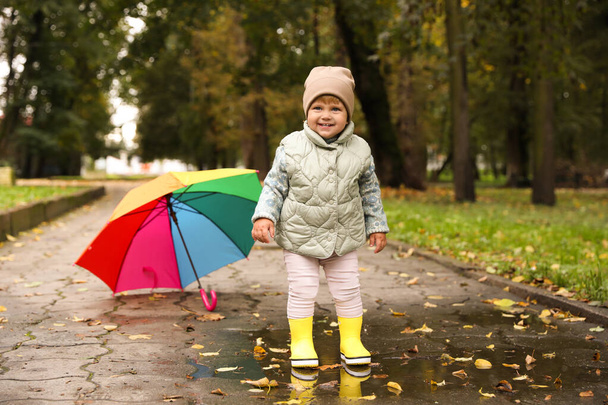 Nettes kleines Mädchen steht in Pfütze neben buntem Regenschirm im Freien - Foto, Bild
