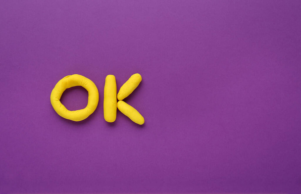 Word Ok made of yellow plasticine on purple background, top view. Space for text - Foto, Imagen