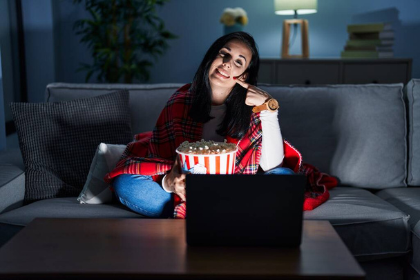 ヒスパニック系の女性は、ソファの上の映画を見てポップコーンを食べて陽気に見て笑って指の歯と口を指しています。歯の健康の概念.  - 写真・画像