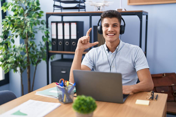 De jonge Spaanse man die op kantoor werkt en een koptelefoon draagt glimlachend en zelfverzekerd gebaar met de hand en een klein bord met vingers en de camera. meetconcept.  - Foto, afbeelding