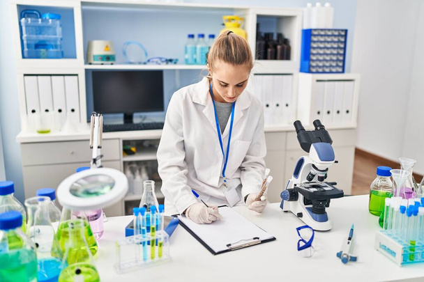 Young blonde woman wearing scientist uniform holding test tubes writing on checklist at laboratory - Foto, immagini