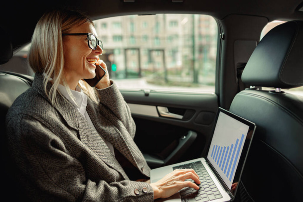 Businesswoman working on laptop and talking phone during way to office in car. High quality photo - Фото, зображення