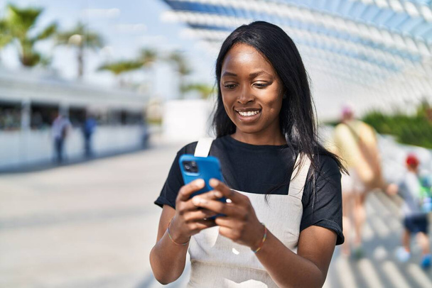 Młoda Afroamerykanka uśmiecha się pewnie używając smartfona w parku - Zdjęcie, obraz