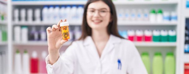 Молодая кавказка-фармацевт улыбается уверенно держа бутылку с таблетками в аптеке - Фото, изображение