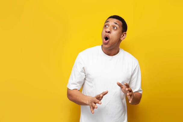 young shocked african american guy in white oversized t-shirt is surprised on yellow isolated background and looks to the side, man in fear looks at the copy space and is surprised - Fotografie, Obrázek