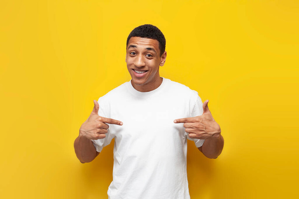 молодой афро-американец в белой огромной футболке показывает свои руки самому себе, мужчина указывает пальцами на грудь на желтом изолированном фоне - Фото, изображение