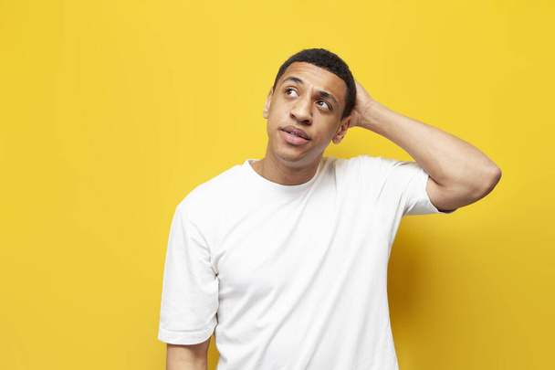young pensive guy afro american confused scratches his head and remembers on yellow isolated background, unsure man is interested in question and looks at copy space - Foto, Bild