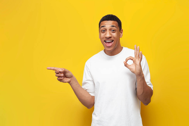 young african american guy in white oversized t-shirt shows gesture ok and advertises copy space, man points his hand to the side on yellow isolated background - 写真・画像