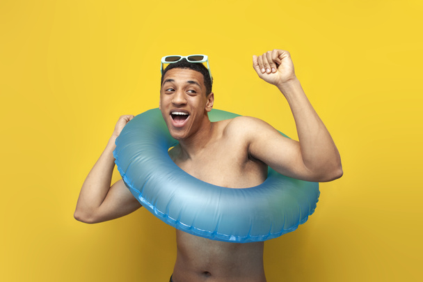 chico afroamericano desnudo alegre en gafas de sol con bailes inflables círculo de natación sobre fondo amarillo, turista masculino en el verano en el mar, concepto de viajes y vacaciones de verano - Foto, imagen