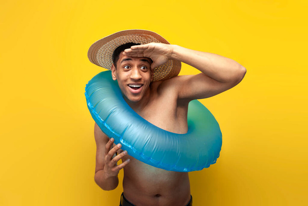 chico afroamericano desnudo alegre en gafas de sol y sombrero con anillo de natación inflable mira en la distancia sobre fondo amarillo, turista masculino en verano, concepto de viaje y vacaciones de verano - Foto, imagen