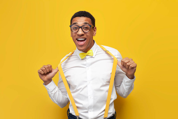 молодий афро-американський хлопець у святковому вбранні з краваткою і підтяжками на жовтому ізольованому фоні, нервовий чоловік в окулярах посміхається і позує
 - Фото, зображення