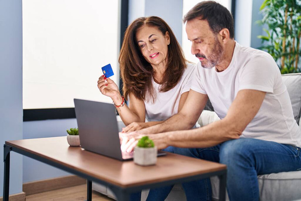 Pareja de hombre y mujer de mediana edad usando computadora portátil y tarjeta de crédito sentada en el sofá en casa - Foto, imagen