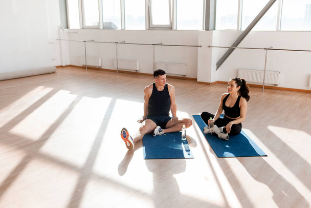joven pareja atlética en ropa deportiva en el entrenamiento están sentados en la estera de yoga y hablando, el hombre y la mujer están haciendo fitness por la mañana y charlando en el descanso - Foto, imagen