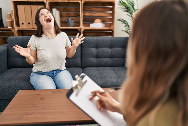Hispanic girl with down syndrome doing therapy crazy and mad shouting and yelling with aggressive expression and arms raised. frustration concept.  - 写真・画像
