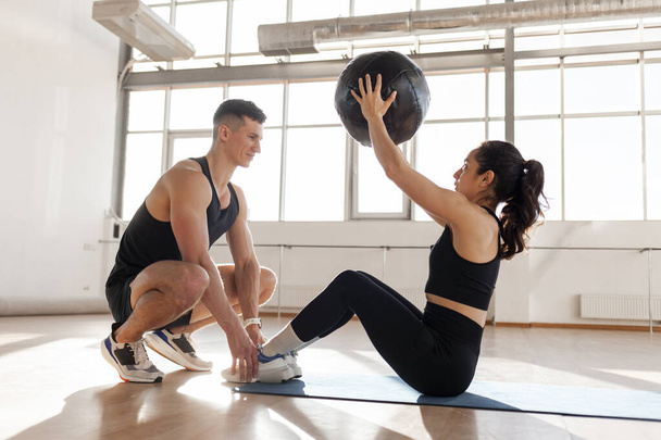 young beautiful athletic couple in sportswear train with ball in fitness room in the morning, fitness girl does exercise on the press with trainer, man trainer helps girl go in for sports - Foto, afbeelding