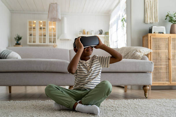 Agradável sorrindo afro-americano estudante medindo metaverso imerso em óculos de toque mundo virtual tentar explorar novos óculos de realidade 3D. Menino despreocupado estudo on-line em fones de ouvido VR em casa - Foto, Imagem