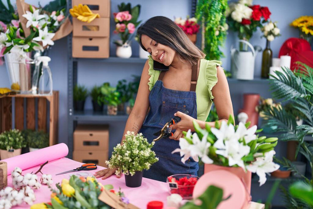 Young hispanic woman florist talking on smartphone cutting plant at florist store - Fotó, kép