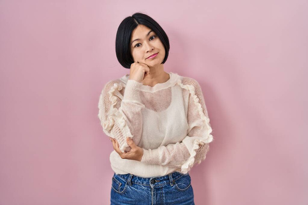 Young asian woman standing over pink background looking confident at the camera with smile with crossed arms and hand raised on chin. thinking positive.  - Foto, Bild
