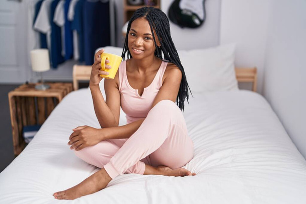 African american woman drinking cup of coffee sitting on bed at bedroom - Foto, imagen