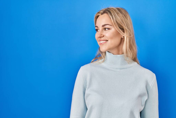 Young caucasian woman standing over blue background looking away to side with smile on face, natural expression. laughing confident.  - Foto, imagen