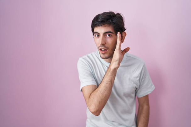 Junger hispanischer Mann steht über rosa Hintergrund Hand auf Mund und erzählt geheime Gerüchte, flüstert bösartige Gespräche  - Foto, Bild