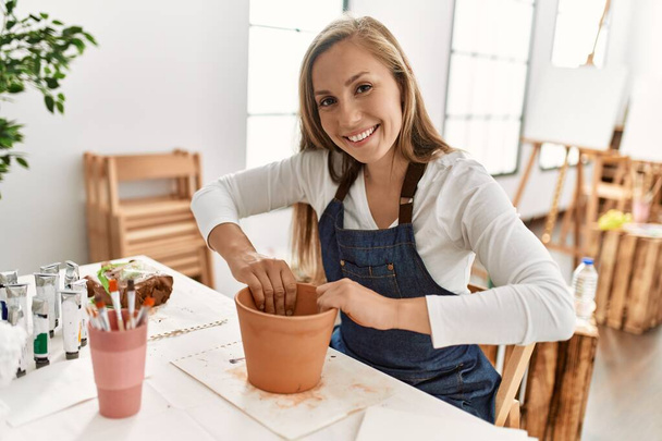 Молодая белая женщина, улыбающаяся уверенно, делает глиняный горшок ручной работы в художественной студии - Фото, изображение