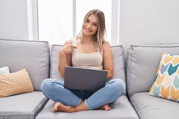 Blonde kaukasische Frau mit Laptop zu Hause sitzt auf dem Sofa und zeigt mit dem Finger auf ein sich selbst lächelndes glücklich und stolz  - Foto, Bild