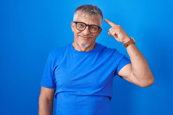 Latinomies harmaat hiukset seisoo sininen tausta hymyillen pään yhdellä sormella, hyvä idea tai ajatus, hyvä muisti  - Valokuva, kuva