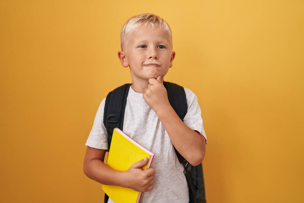 Little caucasian boy wearing student backpack and holding book serious face thinking about question with hand on chin, thoughtful about confusing idea  - Fotografie, Obrázek