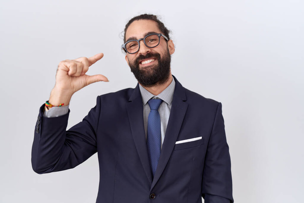 Hispanic man with beard wearing suit and tie smiling and confident gesturing with hand doing small size sign with fingers looking and the camera. measure concept.  - Foto, Imagem