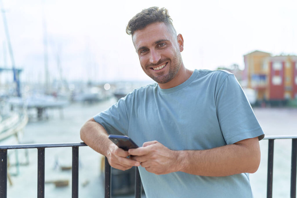 Nuori latino mies hymyilee luottavainen käyttäen älypuhelin meren rannalla - Valokuva, kuva