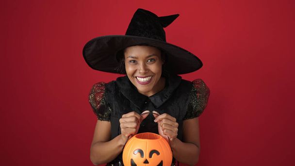 African american woman wearing witch costume holding halloween pumpkin basket over isolated red background - Foto, Imagem