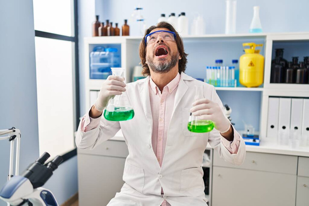 Jóképű középkorú férfi dolgozik tudós laboratóriumban bosszús és frusztrált kiabál a harag, kiabál őrült harag és felemelt kézzel  - Fotó, kép