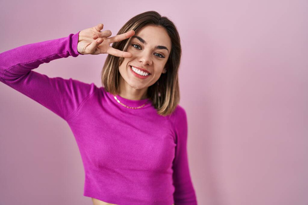 Hispanic woman standing over pink background doing peace symbol with fingers over face, smiling cheerful showing victory  - Fotografie, Obrázek