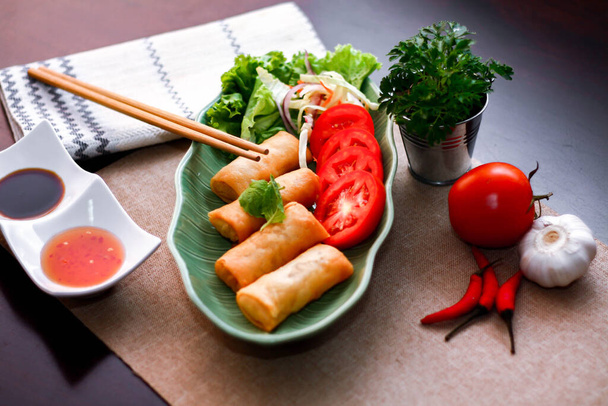 Rollos de primavera fritos, verduras y tomates colocados en un plato de forma de hoja verde sobre una mesa de madera negra y salsa de inmersión en Tailandia. - Foto, Imagen