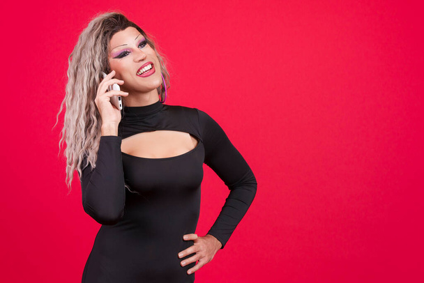 Feliz transexual vestida de drag queen hablando con el móvil en estudio con fondo rojo - Foto, Imagen