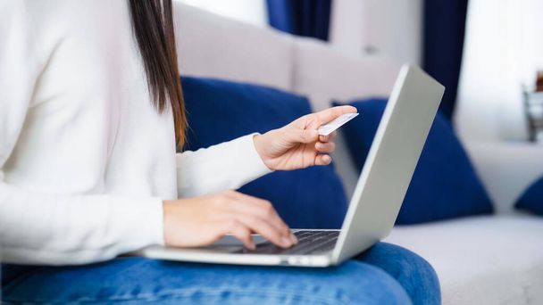 Молода азіатка, сидячи на дивані у вітальні, здійснює онлайн-банківські платежі через Інтернет з банківської картки на комп'ютерному ноутбуці. Шопінг онлайн на ноутбуці з кредитною карткою
 - Фото, зображення