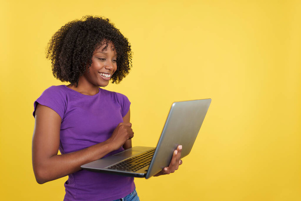 Woman with afro hairstyle smiling while using a laptop in studio with yellow background - Foto, imagen