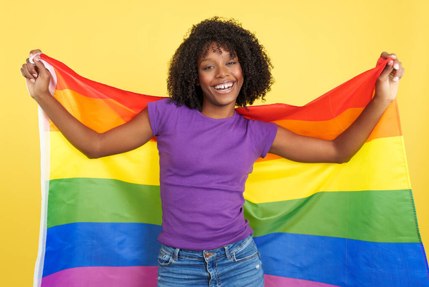 Donna felice con i capelli afro sorridente guardando la fotocamera e alzando una bandiera arcobaleno lgbt in studio con sfondo giallo - Foto, immagini