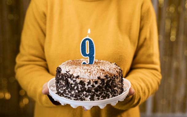 Woman holding birthday cake with burning candles number 9 against decorated background, close up. Celebrates birthday holiday at home. Copy space - 写真・画像